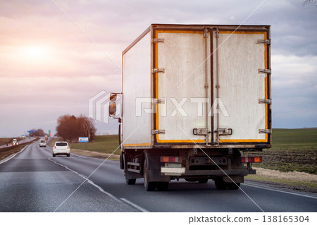 Rear view of cargo truck driving on highway under overcast sky, transportation of goods across landscape, commercial logistics and delivery service, evening sunset. Copy space for text 138165304