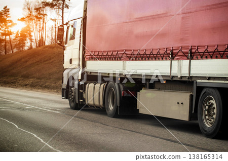 Heavy white cargo truck with red tarp trailer transports goods along asphalt highway, wheels show motion blur, scene features bare trees and cloudy sky 138165314