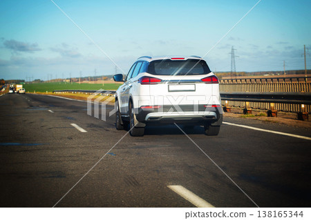 White SUV driving on asphalt highway road under clear blue sky with power lines and green fields in background during bright sunny day journey travel transport concept White SUV driving on asphalt highway road under clear blue sky with power lines and green fields in background during bright sunny day journey travel transport concept 138165344