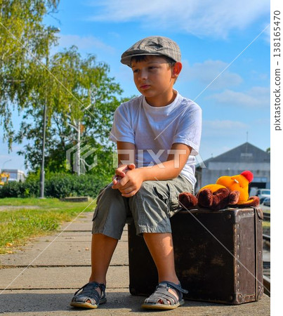 A cute little boy on a train station platform. A sad little boy sits on an old suitcase. The concept of travel, vacation, and a carefree childhood. Retro and vintage vibe. 138165470