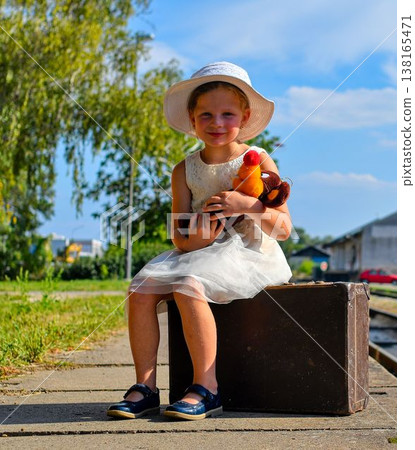A lovely girl at the train station. A charming girl at the train station. A little girl holds a cuddly toy and sits on an old suitcase. The concept of travel, summer vacation, and a carefree childhood A lovely girl at the train station. A charming girl at the train station. A little girl holds a cuddly toy and sits on an old suitcase. The concept of travel, summer vacation, and a carefree childhood 138165471
