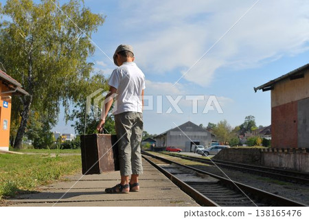 A cute little boy on a train station platform. A sad little boy in a flat cap with an old suitcase, leaves the railway platform. The concept of travel, holidays, and a carefree childhood. Retro and A cute little boy on a train station platform. A sad little boy in a flat cap with an old suitcase, leaves the railway platform. The concept of travel, holidays, and a carefree childhood. Retro and 138165476