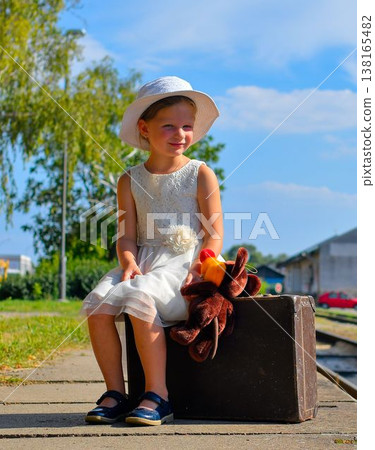A cute little girl in a white dress with a hat and a soft toy sits on an old suitcase. A cutie with a smile on a train platform. The concept of summer vacation, travel, and a carefree childhood and 138165482