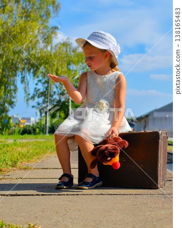 A lovely little girl in a white dress with a cuddly toy sits on an old suitcase. A sweet smiling girl on a train platform. The concept of summer holidays, travel, and a carefree childhood and family. 138165483