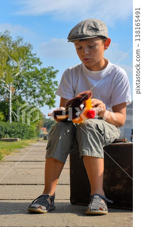 Cute boy at the train station. A sad little boy holds a stuffed toy and sits on an old suitcase. The concept of travel, vacation, and carefree childhood. Retro and vintage vibe. Cute boy at the train station. A sad little boy holds a stuffed toy and sits on an old suitcase. The concept of travel, vacation, and carefree childhood. Retro and vintage vibe. 138165491
