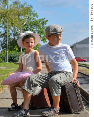 Lovely siblings on a train platform. A small girl and boy are traveling on vacation. The concept of travel, holidays, and a carefree childhood. Vintage and retro mood. Lovely siblings on a train platform. A small girl and boy are traveling on vacation. The concept of travel, holidays, and a carefree childhood. Vintage and retro mood. 138165495