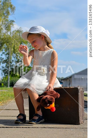 A cute small girl in a white dress with a plush toy and an old suitcase. The adorable smiling girl is standing on a train platform. The concept of the end of summer vacation, traveling, and a carefree A cute small girl in a white dress with a plush toy and an old suitcase. The adorable smiling girl is standing on a train platform. The concept of the end of summer vacation, traveling, and a carefree 138165496