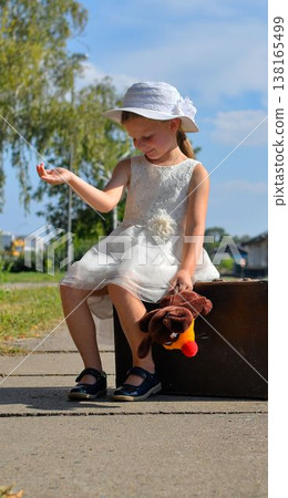 A cute small girl in a white dress with a plush toy and an old suitcase. The adorable smiling girl is standing on a train platform. The concept of the end of summer vacation, traveling, and a carefree A cute small girl in a white dress with a plush toy and an old suitcase. The adorable smiling girl is standing on a train platform. The concept of the end of summer vacation, traveling, and a carefree 138165499