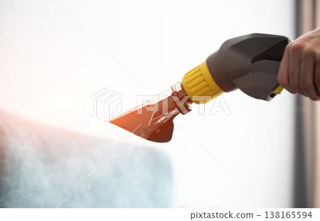 One person hand operating cleaning machine nozzle applying spray of water and detergent on textile surface for deep cleaning upholstery furniture indoors 138165594