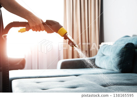 Person hand holds professional cleaning machine nozzle spraying water solution onto blue sectional sofa upholstery for deep cleaning service indoors 138165595