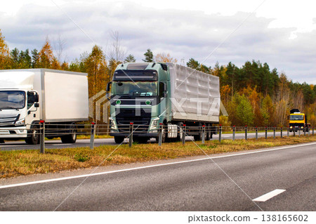 Green semitrailer truck with cargo moving on highway road. White box truck and yellow heavy vehicle on autumn forest background. Freight transport. Logistics. Delivery of goods. 138165602