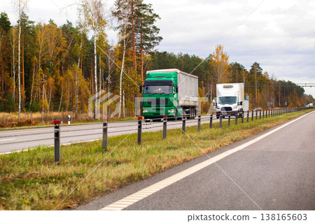 Green semi-truck and white delivery truck on multilane highway. Cargo transportation on autumn road through forest under cloudy sky. Commercial vehicle movement. Green semi-truck and white delivery truck on multilane highway. Cargo transportation on autumn road through forest under cloudy sky. Commercial vehicle movement. 138165603
