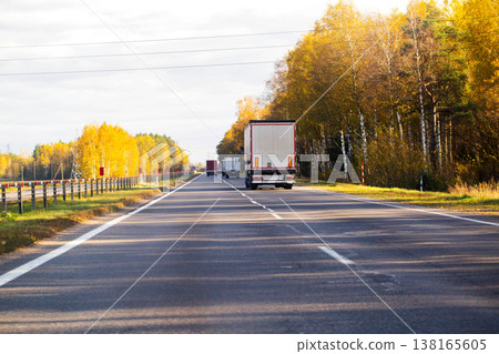 Highway view with line of trucks driving away from camera, bordered by vibrant autumn forest foliage under clear sky with overhead power lines. Highway view with line of trucks driving away from camera, bordered by vibrant autumn forest foliage under clear sky with overhead power lines. 138165605