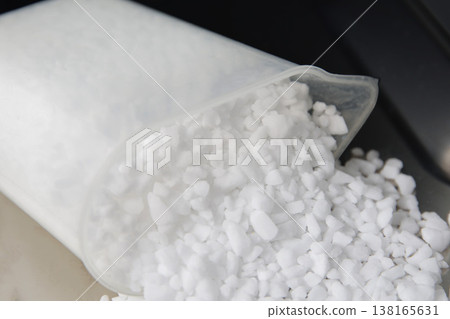 White granular dishwasher salt spilling from translucent plastic scoop onto light surface. Focus on irregular crystals, showing rough texture and purity of household product. 138165631