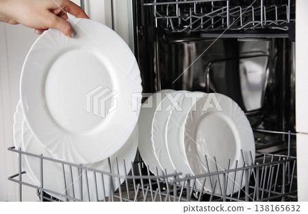 Human hand placing collection of white elegant ceramic plates onto lower rack of contemporary built-in dishwasher, showcasing modern kitchen chore. 138165632