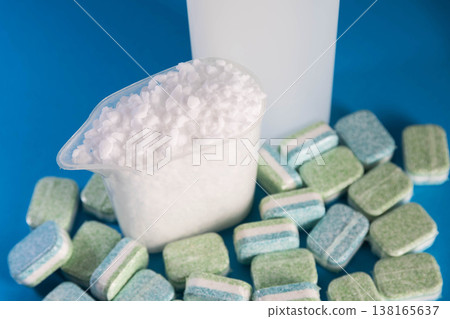 Dishwasher salt in measuring cup with rinse aid bottle and numerous detergent tablets for kitchen appliance cleaning and maintenance on blue background Dishwasher salt in measuring cup with rinse aid bottle and numerous detergent tablets for kitchen appliance cleaning and maintenance on blue background 138165637