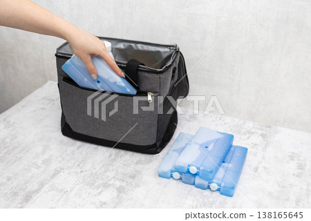 Woman hand placing blue rectangular ice pack inside open grey fabric cooler bag. Multiple cold generators next to bag on light textured surface, preparation for travel or picnic. 138165645