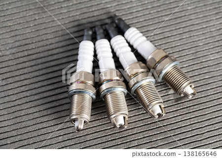 Four new vehicle engine spark plugs with white ceramic insulators and metallic bodies on dark textured surface for automotive service. Four new vehicle engine spark plugs with white ceramic insulators and metallic bodies on dark textured surface for automotive service. 138165646