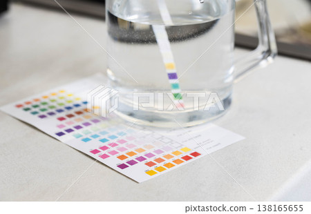 Water quality test strip measurement of parameters in glass pitcher with clear liquid, comparing results with color chart for accurate analysis of water composition. 138165655