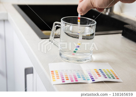 Hand holds water test strip dipping into glass mug with water on kitchen counter next to color chart for measurement of water quality parameters, home water testing process Hand holds water test strip dipping into glass mug with water on kitchen counter next to color chart for measurement of water quality parameters, home water testing process 138165659