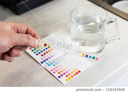 Hand holding water quality test strip displaying colored chemical indicators for comparison with reference chart, transparent glass mug of water next to paper results table 138165660