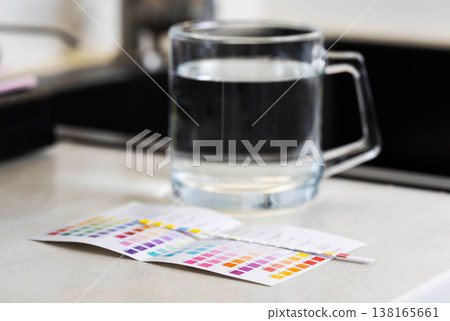 Clear glass mug filled with water, beside it water quality test strip displays results against vibrant color chart on bright countertop, indicating chemical parameter measurement of liquid purity 138165661