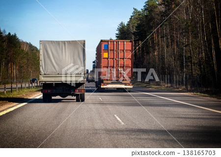Two heavy transport trucks traveling on multi-lane asphalt highway through green pine tree forest under clear blue sky day movement logistic cargo 138165703