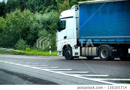 White semi-truck and blue cargo trailer traveling on asphalt highway through green natural landscape with road signs showing speed regulation. Copy space for text White semi-truck and blue cargo trailer traveling on asphalt highway through green natural landscape with road signs showing speed regulation. Copy space for text 138165714