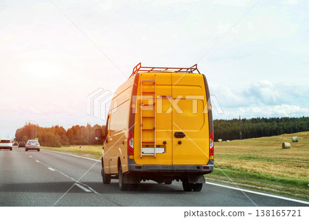 Yellow cargo van traveling on highway for commercial transport of goods, featuring roof rack and ladder on rear, passing agricultural fields with hay bales. Copy space for text Yellow cargo van traveling on highway for commercial transport of goods, featuring roof rack and ladder on rear, passing agricultural fields with hay bales. Copy space for text 138165721