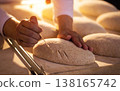 Close-up of artisan hand scoring raw bread dough with sharp tool, preparing multiple rustic loaves for baking process in traditional bakery setting 138165742
