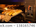 Baker placing fresh golden brown baguettes on metal tray into hot commercial oven for baking process in bakery kitchen industrial setting 138165746