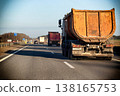 Large orange dump truck traveling on multi-lane highway, commercial transportation vehicle on asphalt road under clear blue sky in rural landscape 138165753