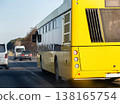 Yellow passenger bus travels on highway road, rear view of vehicle in motion, traffic flow with other cars on sunny day, outdoor transportation scene. 138165754
