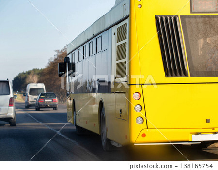 Yellow passenger bus travels on highway road, rear view of vehicle in motion, traffic flow with other cars on sunny day, outdoor transportation scene. 138165754