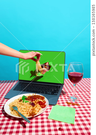 Hand feeding spaghetti into mouth emerging from laptop screen on table with wine. Concept of food delivery platform, digital consumption, online service advertising. Hand feeding spaghetti into mouth emerging from laptop screen on table with wine. Concept of food delivery platform, digital consumption, online service advertising. 138165985
