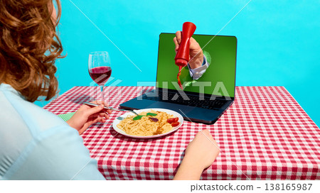 Hand squeezing ketchup from laptop screen onto pasta plate on checkered table with wine glass. Hand squeezing ketchup from laptop screen onto pasta plate on checkered table with wine glass. 138165987