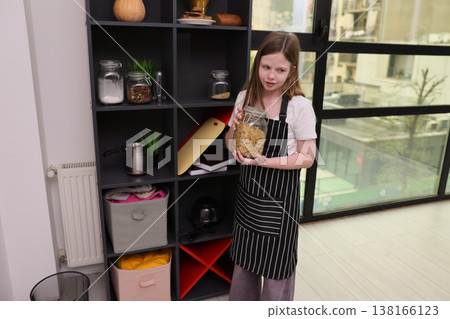 Girl in a striped apron holds a jar of cookies while standing in a cozy kitchen on a sunny day Girl in a striped apron holds a jar of cookies while standing in a cozy kitchen on a sunny day 138166123