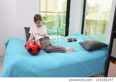 Young boy plays with a bright red soccer ball on a cozy blue bed while looking out at a vibrant city view 138166147
