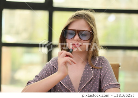 Young girl in stylish sunglasses thinking deeply while holding a lipstick tube in a bright, modern space 138166255