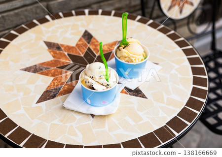 Close up of appetizing ice cream scoops in paper cups resting on a colorful mosaic table at an outdoor cafe. Detailed texture of frozen dessert and vibrant ceramic tiles 138166669