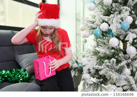 Child in Christmas hat shows mixed emotions while unwrapping a pink gift beside a festive tree full of decorations Child in Christmas hat shows mixed emotions while unwrapping a pink gift beside a festive tree full of decorations 138166696