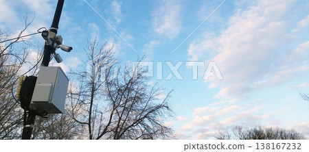 A high pole holds multiple white surveillance cameras and a grey electrical box. Bare trees are set against a blue sky with soft white clouds, symbolizing constant observation in a public space 138167232