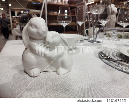 Charming white ceramic figurine of two hugging rabbits rests on linen tablecloth. The glossy sculpture is part of an elegant table setting featuring glassware, plates in brightly lit interior. Easter Charming white ceramic figurine of two hugging rabbits rests on linen tablecloth. The glossy sculpture is part of an elegant table setting featuring glassware, plates in brightly lit interior. Easter 138167235