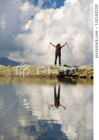 A hiker stands triumphantly on a rocky outcrop overlooking a calm lake. The sky is filled with fluffy clouds, creating a stunning backdrop for this adventurous moment in the Swizz mountains. 138168208