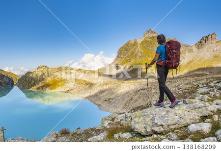 A person stands on rocky terrain, gazing at a stunning turquoise mountain lake surrounded. Dressed in hiking gear and holding trekking poles. Wildsee Pizol, Switzerland. 138168209