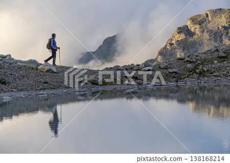 A person stands on rocky terrain, gazing at a stunning turquoise mountain lake surrounded. Dressed in hiking gear and holding trekking poles. Wildsee Pizol, Switzerland. 138168214