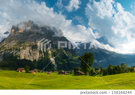 Majestic mountains rise in the background as quaint wooden cabins dot green landscape. A winding path guides a visitor through this serene Swiss alps. Grindelwald Jungfrau Bernese alps Switzerland. Majestic mountains rise in the background as quaint wooden cabins dot green landscape. A winding path guides a visitor through this serene Swiss alps. Grindelwald Jungfrau Bernese alps Switzerland. 138168244