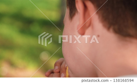 unhealthy food for children concept, cute schoolboy eating burger in garden, child holding burger and taking bite in green garden, close portrait of schoolboy eating fast food, kid eating cheeseburger 138168594