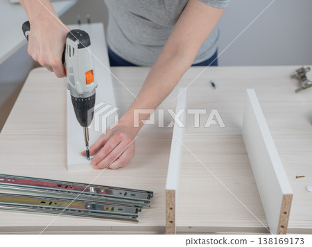 A Caucasian woman uses a screwdriver to tighten a screw while assembling a cabinet. A Caucasian woman uses a screwdriver to tighten a screw while assembling a cabinet. 138169173