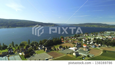 View of a lake surrounded by hills and farms during a sunny day with boats on the water and houses along the shore View of a lake surrounded by hills and farms during a sunny day with boats on the water and houses along the shore 138169786